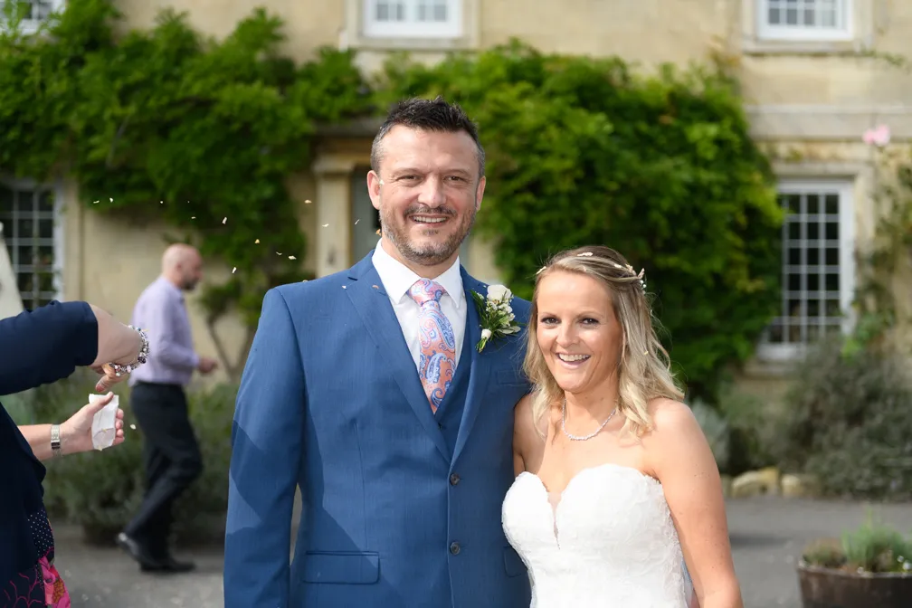 Bride in a strapless white wedding dress and groom in a blue suit with a pink paisley tie smiling outdoors in front of a building with greenery.