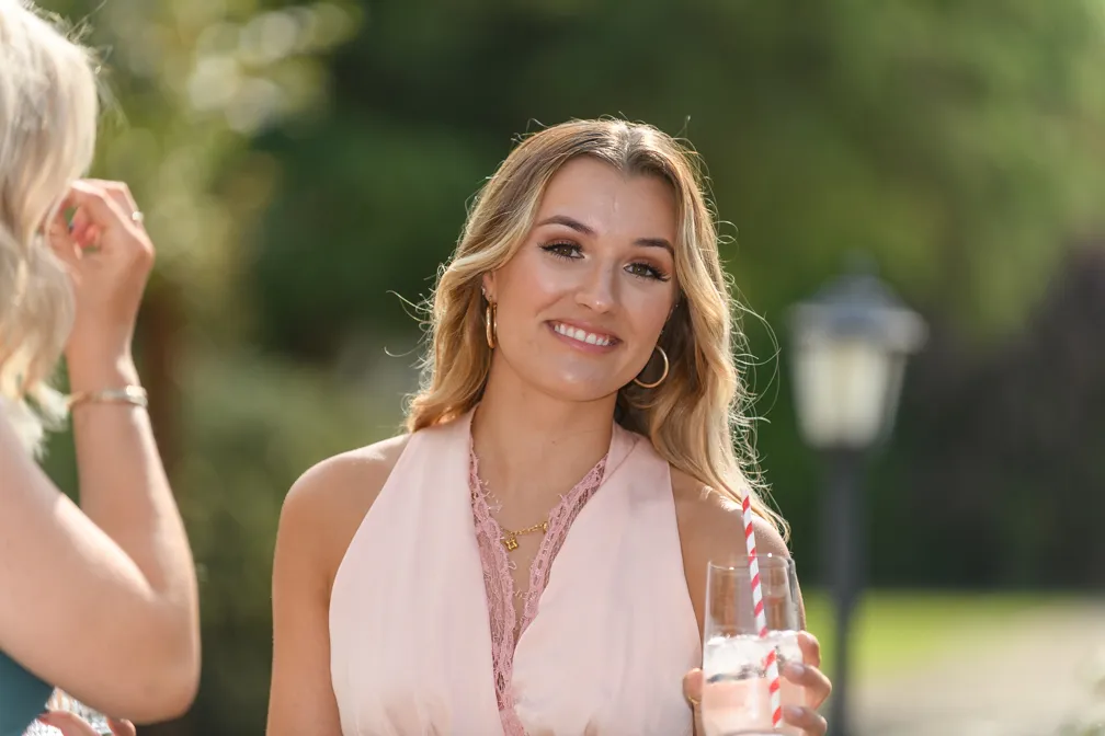 Smiling woman in a light pink dress holding a glass of water with a red and white striped straw outdoors.