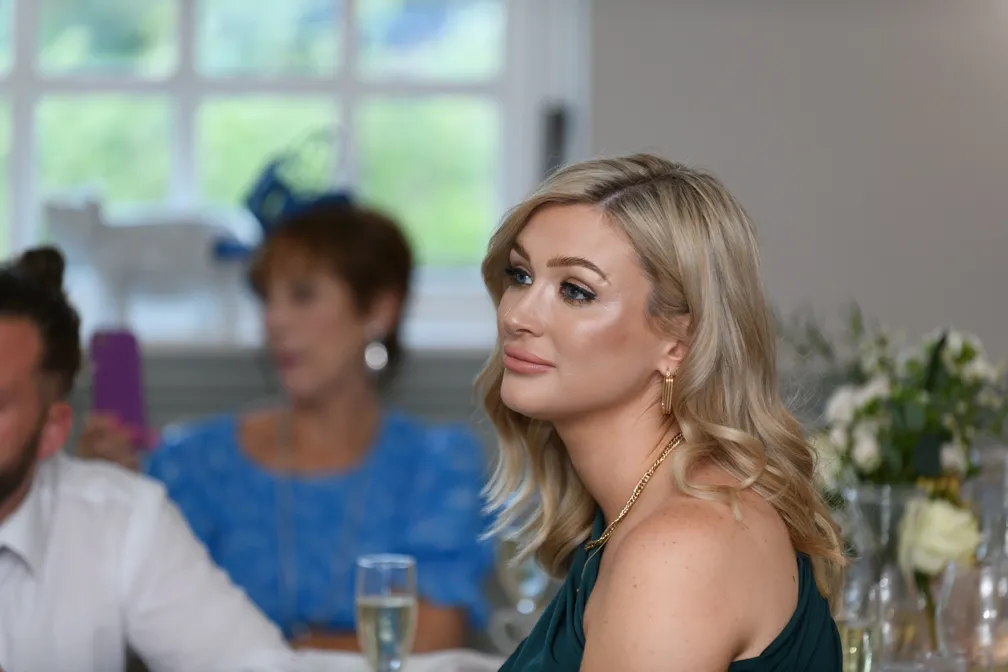 Blonde woman in a dark green dress with gold jewelry looking thoughtfully to the side at an indoor event.