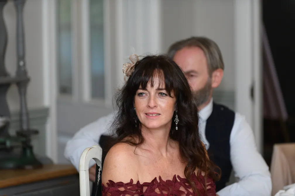 Woman with long dark hair wearing a burgundy off-shoulder dress and dangling earrings seated indoors with a man blurred in the background.
