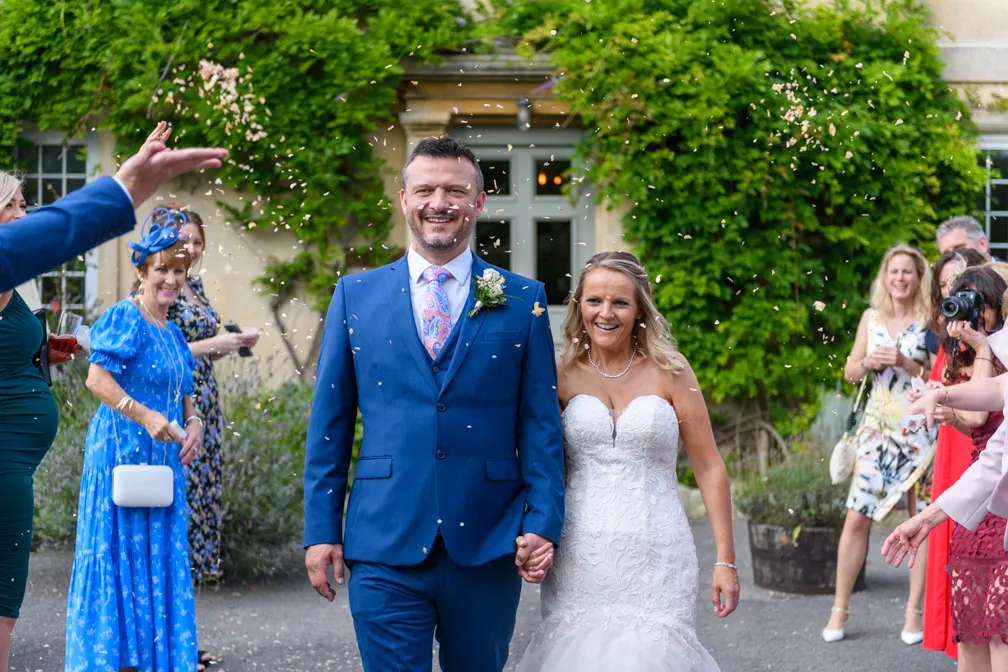 Smiling bride in white lace gown and groom in blue suit holding hands while guests throw confetti outdoors.