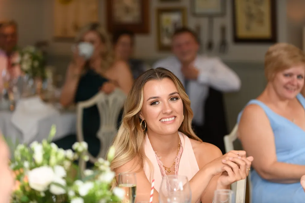 Smiling woman with long blonde hair wearing a light pink dress sitting at a table with blurred people and floral centerpiece in the background.