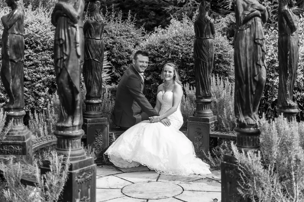 Bride and groom holding hands and sitting on a bench surrounded by statues in a garden.