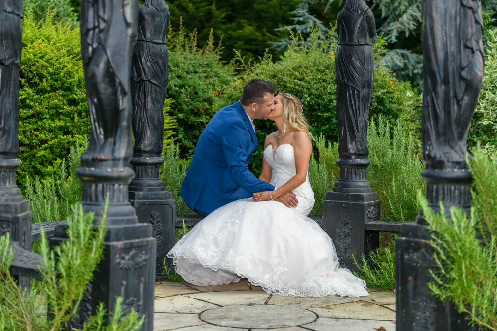 Bride in white wedding dress and groom in blue suit sitting and kissing in a garden gazebo surrounded by greenery.