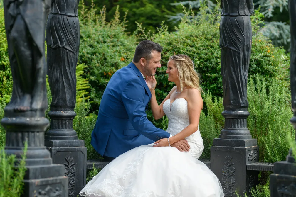 Bride in a white lace gown and groom in a blue suit sitting closely and affectionately facing each other amid garden greenery and black statues.