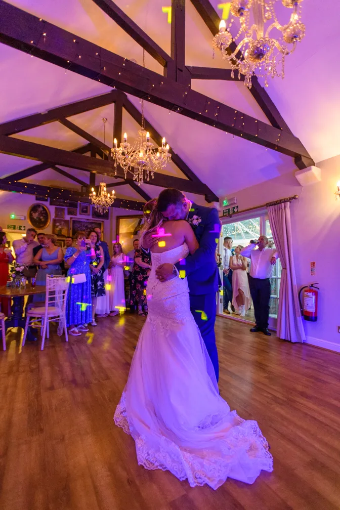 Bride and groom embrace while sharing a first dance in a warmly lit wooden-beamed reception hall with guests watching.