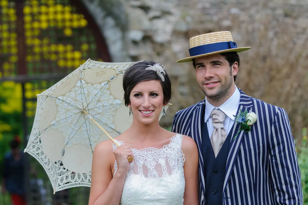 Bride in lace dress holding a lace parasol next to groom in striped suit and straw boater hat.