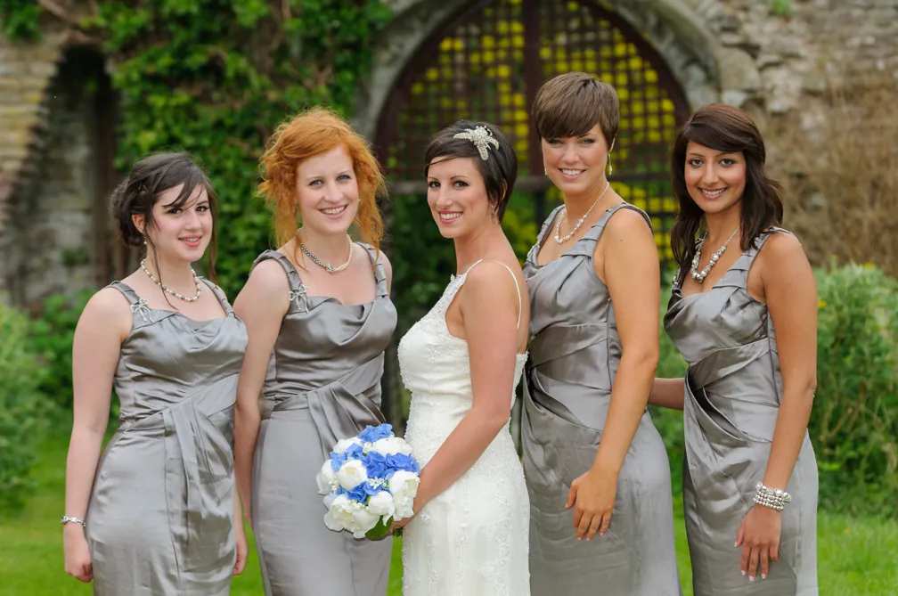 Bride in white dress holding a blue and white bouquet standing with four bridesmaids wearing silver-gray dresses outdoors.