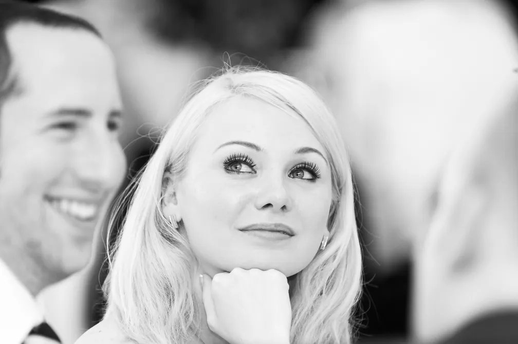 Black and white close-up of a blonde woman looking up thoughtfully with her chin resting on her hand, two blurred men on either side.