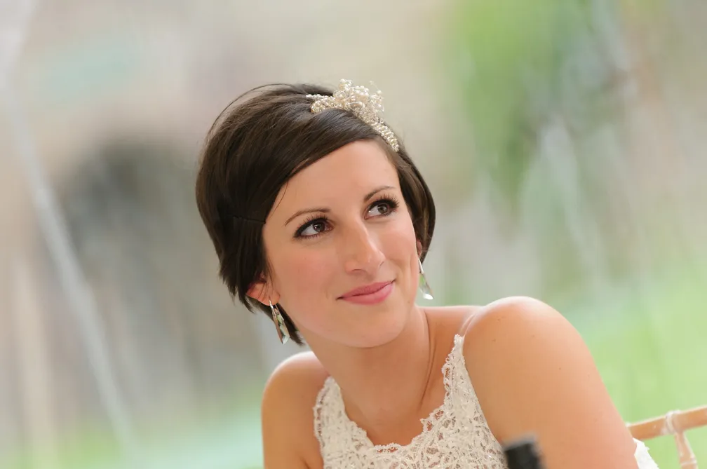 Smiling woman with short dark hair wearing a white lace top and jeweled hair accessory.
