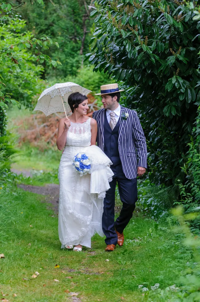 Bride in white lace dress holding a bouquet and umbrella walking arm-in-arm with groom wearing a striped jacket and straw boater hat in a lush green garden.