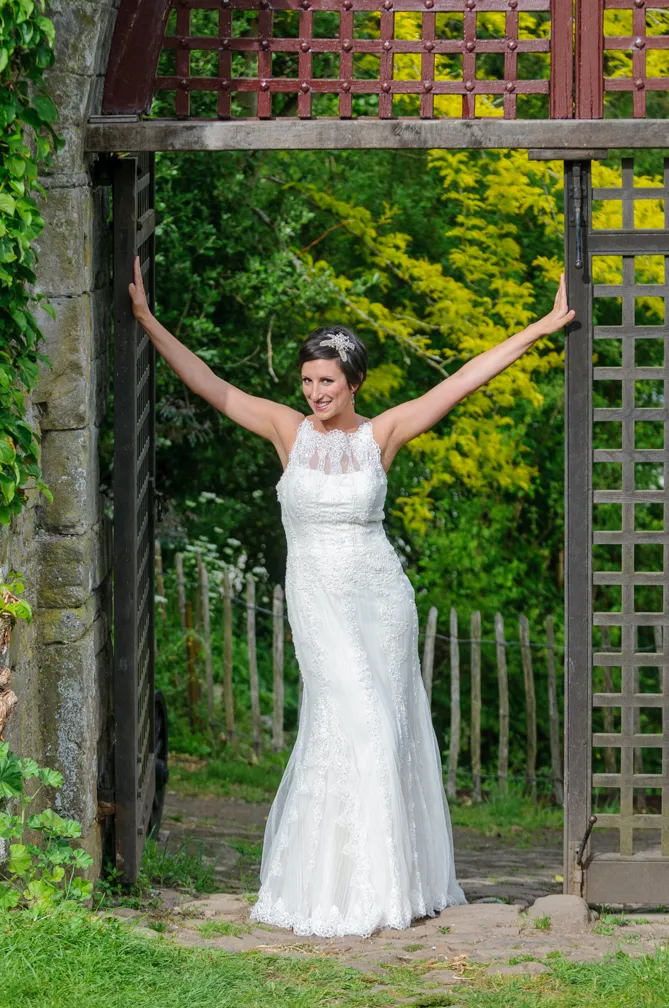 Smiling bride in a white lace wedding dress standing under a rustic wooden gate with greenery and a wooden fence in the background.