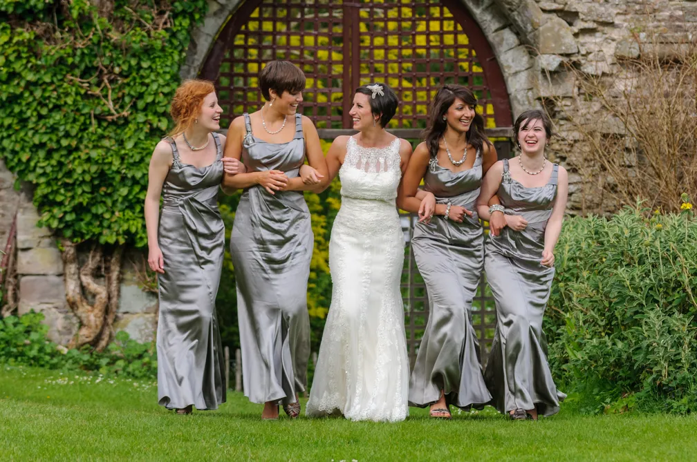 Bride in white lace dress walking arm-in-arm with four bridesmaids wearing matching silver satin gowns outdoors in front of a stone archway with greenery.