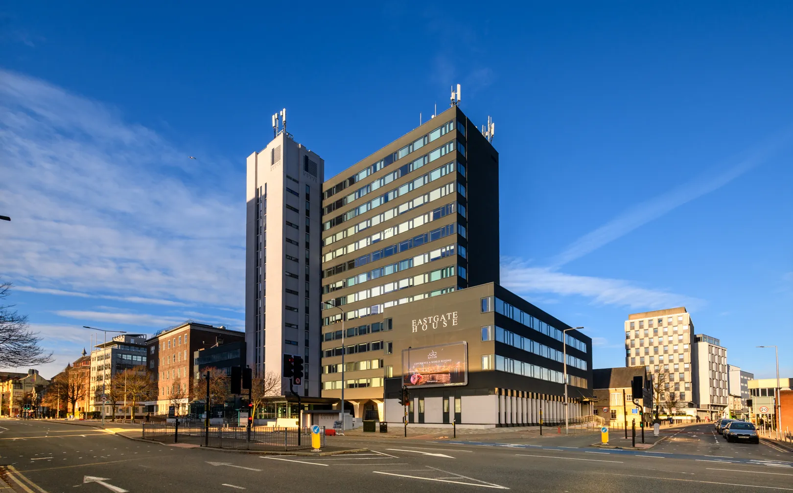 Eastgate House office building under blue sky at a city street intersection with traffic lights and parked cars.