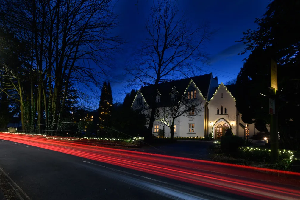 White house decorated with white string lights at night with red light trails from passing cars on the road in front.