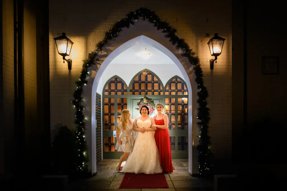 Bride in white gown smiling between two bridesmaids in red and champagne dresses under a decorated archway at night.
