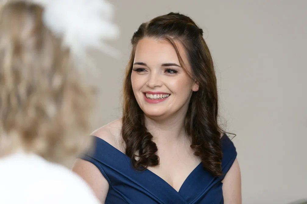 Smiling woman with brown curled hair wearing a navy blue off-shoulder dress, looking to the side.