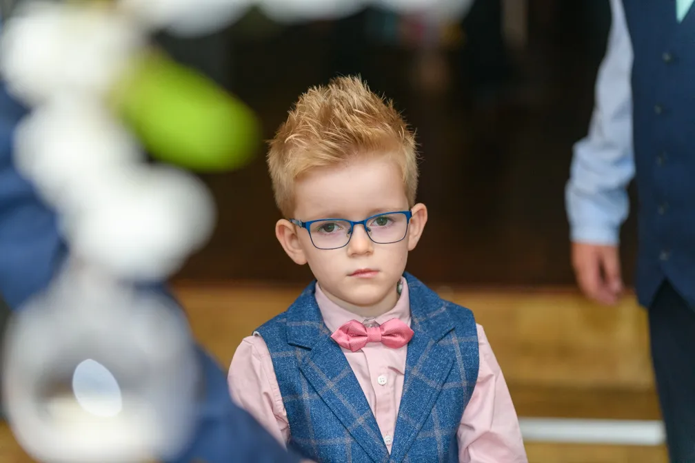 Young boy with spiked blonde hair wearing blue glasses, pink shirt, blue checked vest, and pink bow tie.
