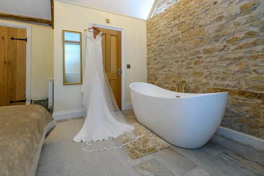 White wedding dress hanging on a wooden door in a room with a white freestanding bathtub and a stone wall.