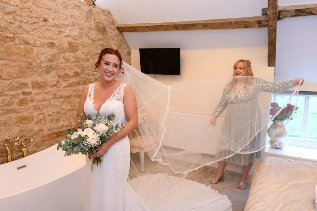 Smiling bride in a white dress holding a bouquet with another woman holding the bride’s long veil in a room with stone wall and window.