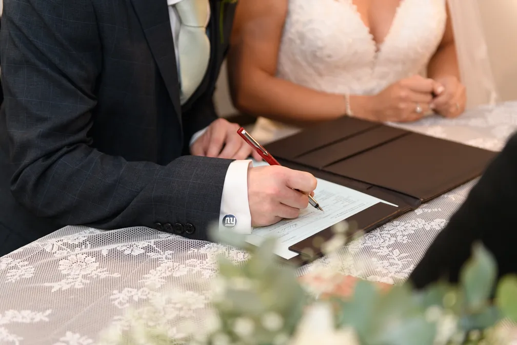 Man in a suit signing a document on a lace-covered table, with a woman in a white wedding dress beside him.