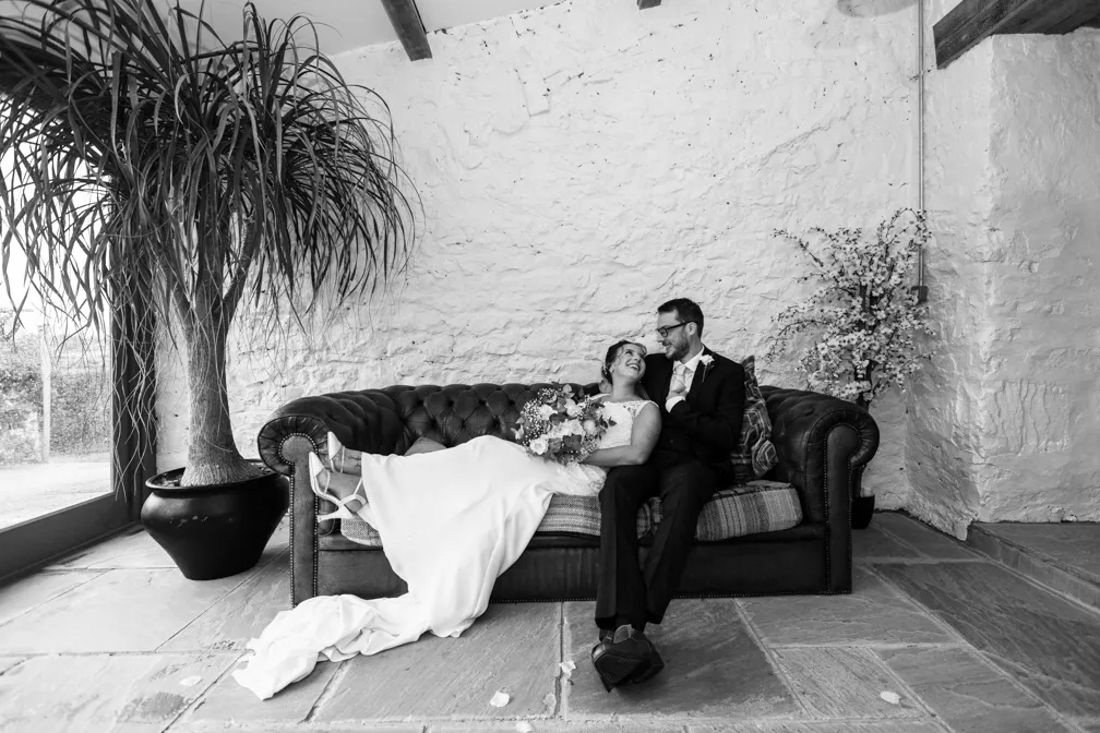 Bride and groom sitting on a leather couch in a rustic room, the bride holding a bouquet and both smiling at each other.
