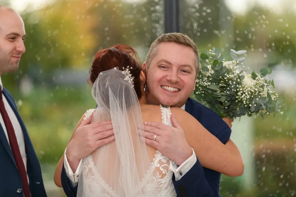 Groom in blue suit smiling and hugging bride in lace dress and veil holding a bouquet, with a man in a suit nearby.