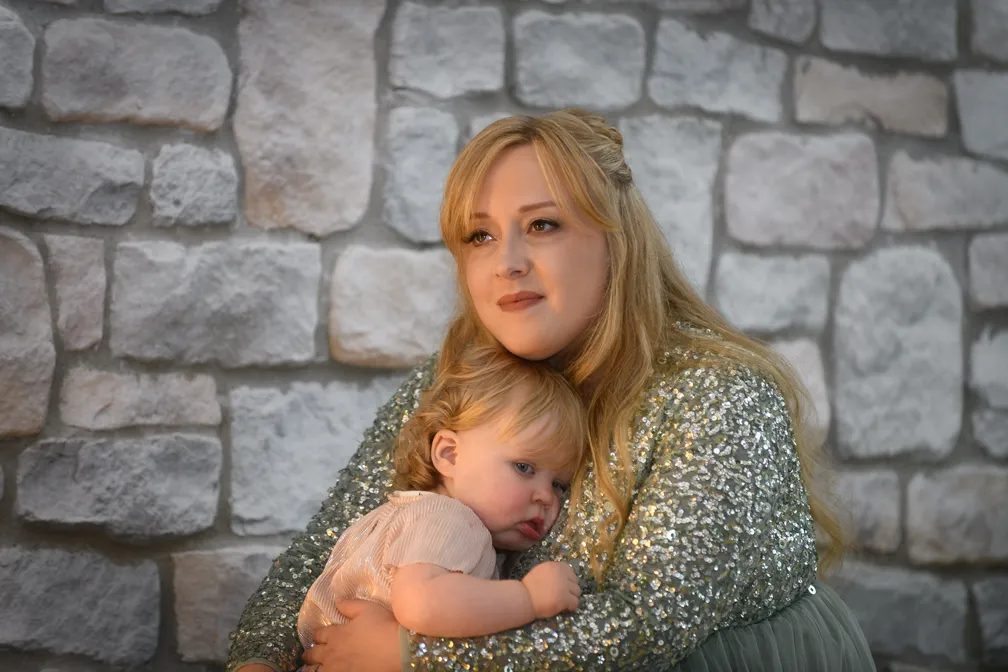 Woman with long blonde hair in a sequined top holding a young child resting on her shoulder against a stone wall background.