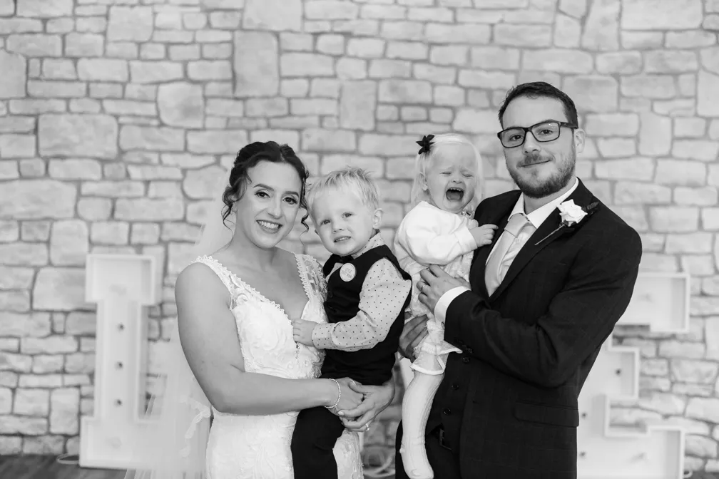 Bride in white lace wedding dress holding a boy and groom in a dark suit holding a crying girl, standing in front of a stone wall with large decorative letters.