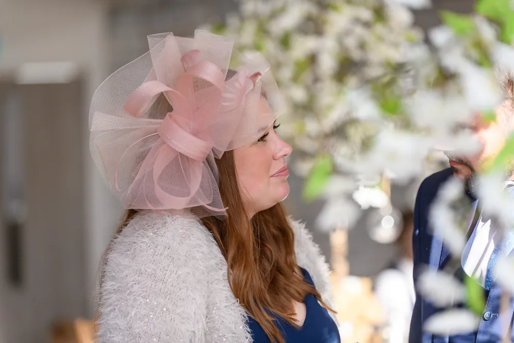 Woman with long brown hair wearing a large pink tulle hat and a fuzzy white sweater looking at a man in a navy suit partially visible behind white floral decorations.