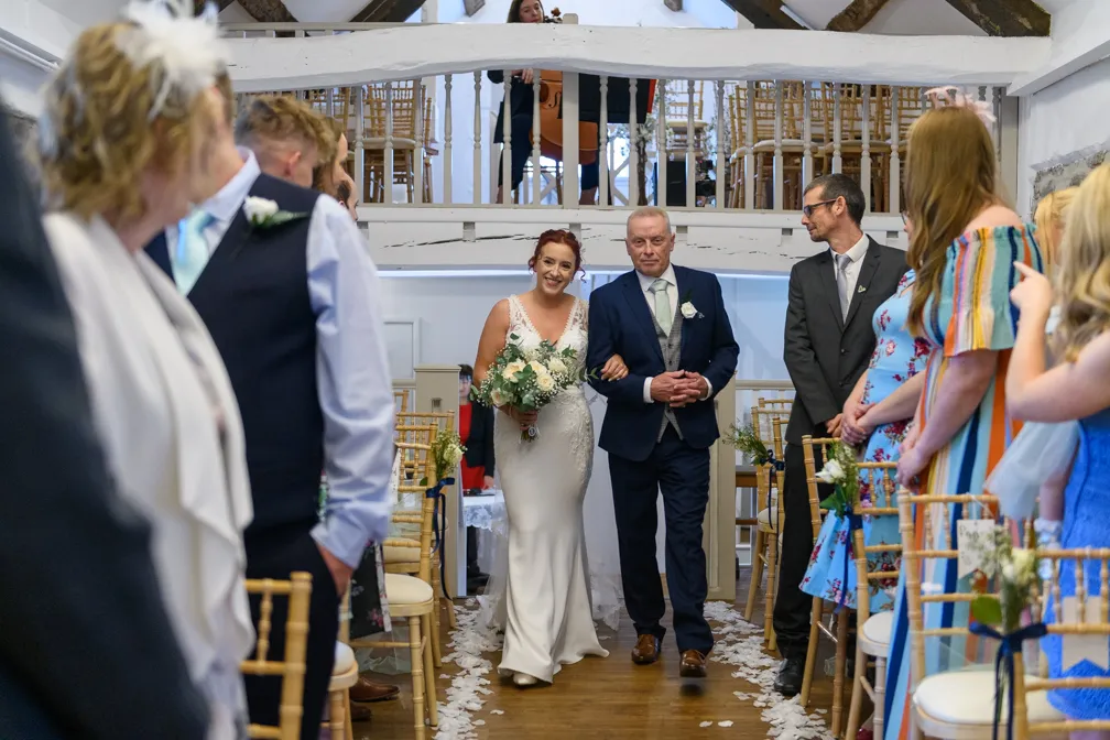 Bride in a white gown holding a bouquet walking arm-in-arm down an aisle lined with seated guests in a rustic indoor venue.