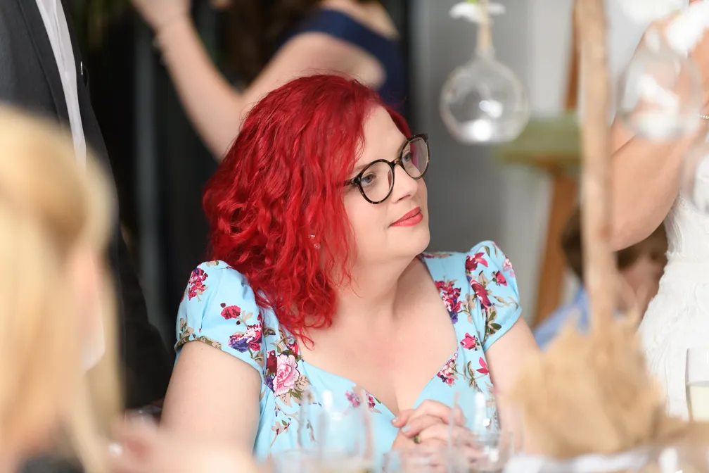 Woman with bright red curly hair and glasses wearing a light blue floral dress sitting at a table.