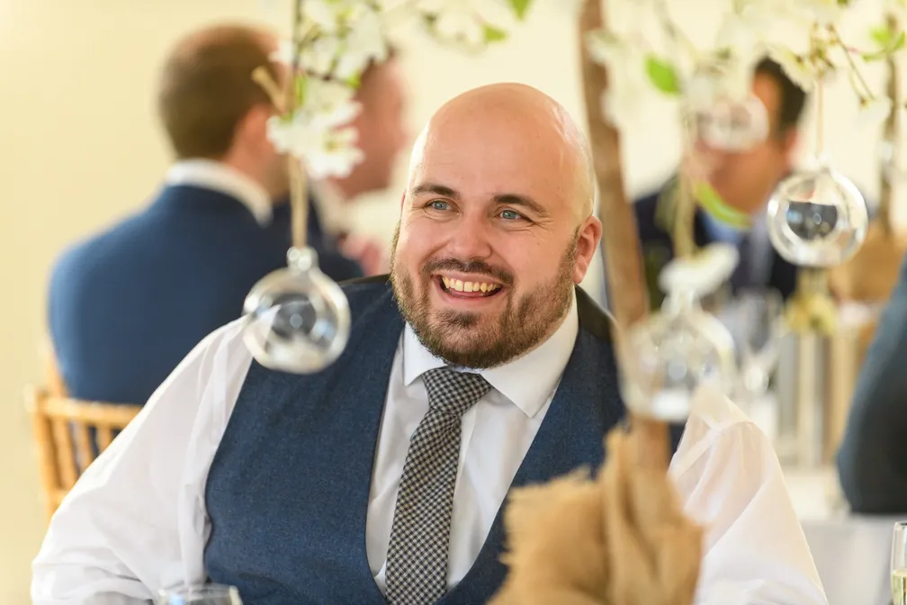 Smiling bald man with a beard wearing a blue vest, white shirt, and checkered tie at a formal event.