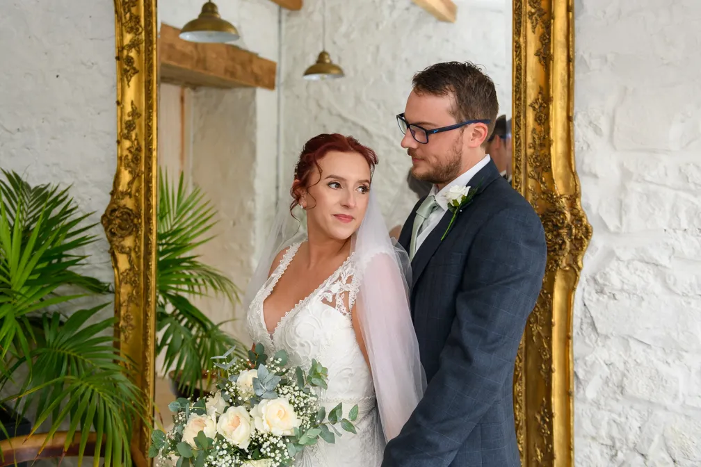 Bride in white lace wedding dress holding a bouquet of white roses and eucalyptus, embraced from behind by groom in blue suit with white boutonniere, standing in front of a large ornate gold-framed mirror and green plants.