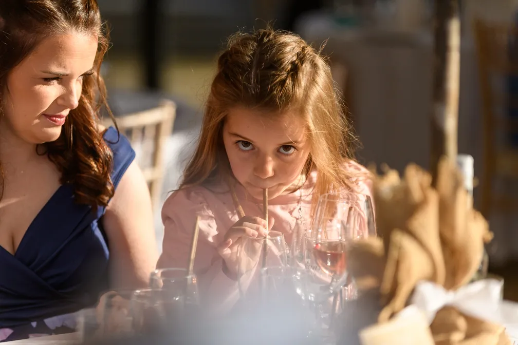 Young girl sipping a drink through a straw at a dining table with a woman beside her.