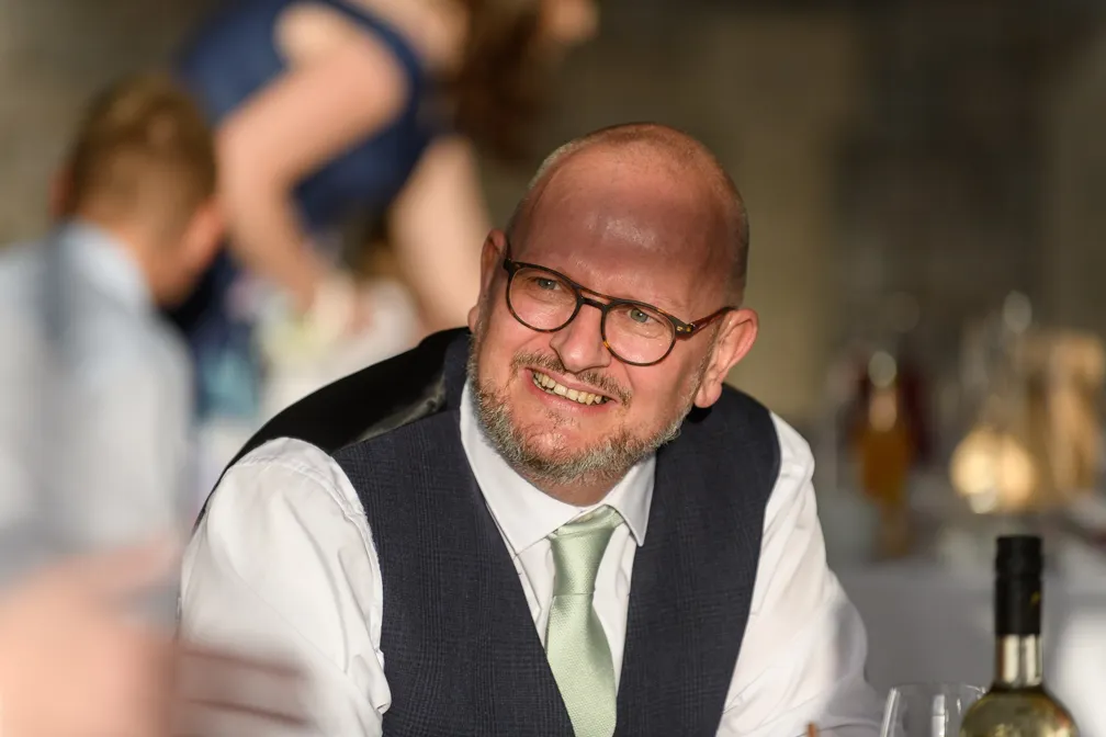 Smiling bald man with beard and glasses wearing a white shirt, light green tie, and dark vest sitting at a table.