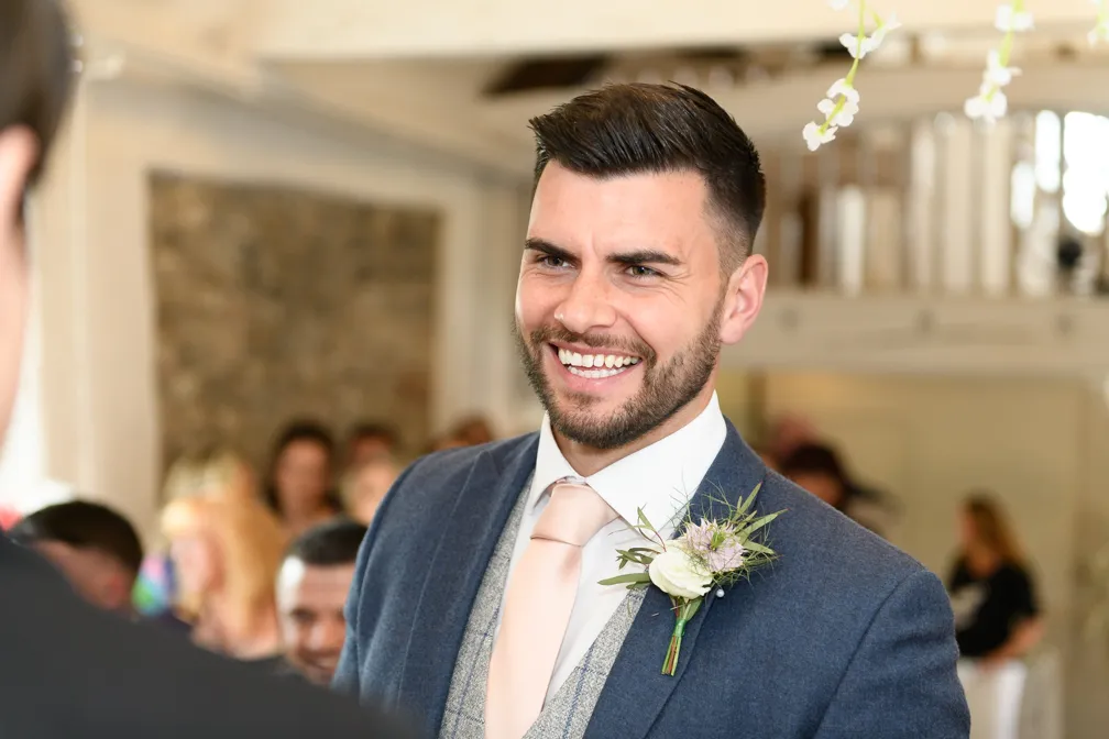Smiling groom in a blue suit with a light pink tie and boutonniere, standing indoors with blurred guests in the background.