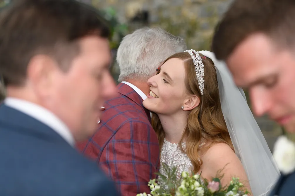 Bride smiling and hugging an older man in a red plaid jacket during a wedding ceremony.