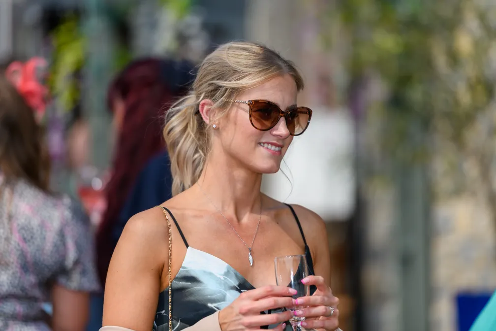 Smiling woman with blonde hair tied back wearing sunglasses and a floral dress, holding a clear glass in both hands.