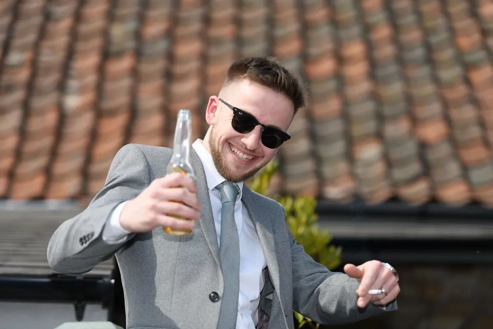 Smiling young man wearing sunglasses, gray suit, and tie, holding a bottle of beer and gesturing with his other hand outdoors.