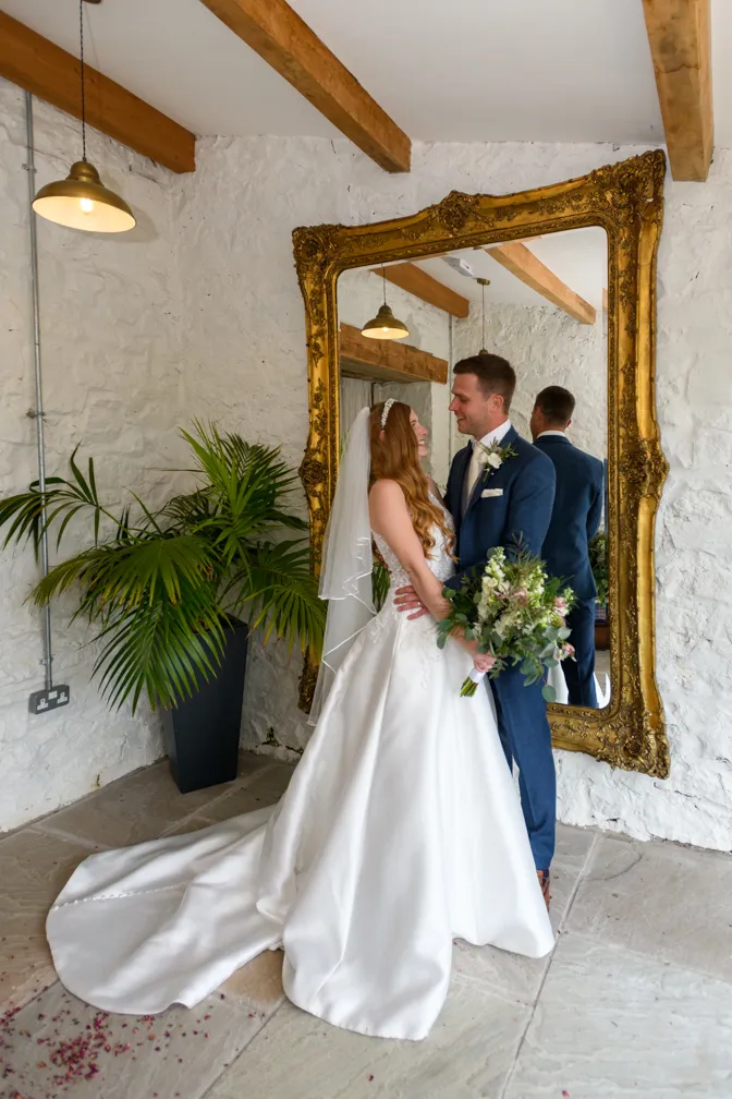 Bride in a white wedding gown holding a bouquet and groom in a blue suit standing close facing each other in front of a large ornate gold-framed mirror.