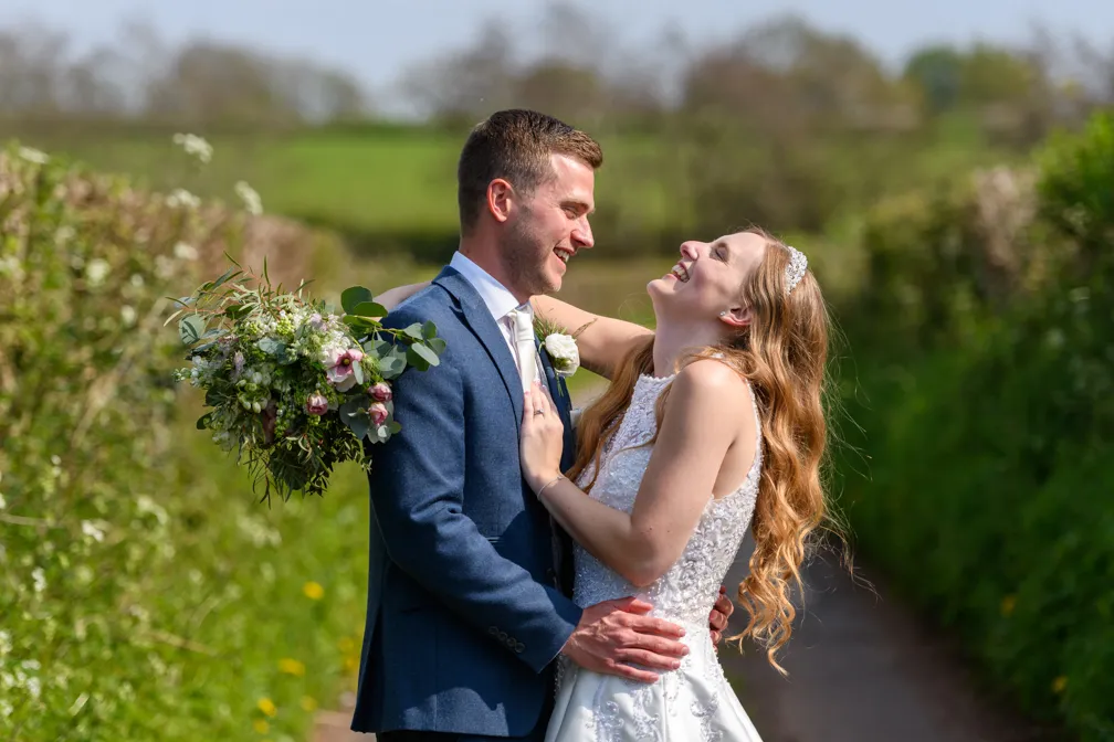 Bride and groom laughing and embracing outdoors with green hedges in the background.