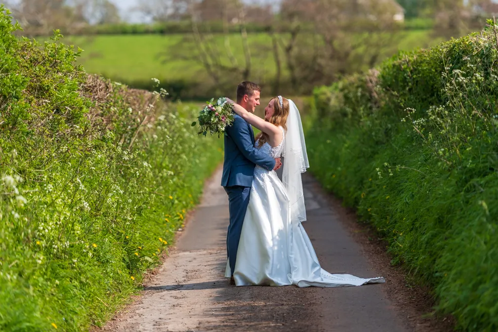 Bride and groom embracing on a country road lined with green hedges and grass.