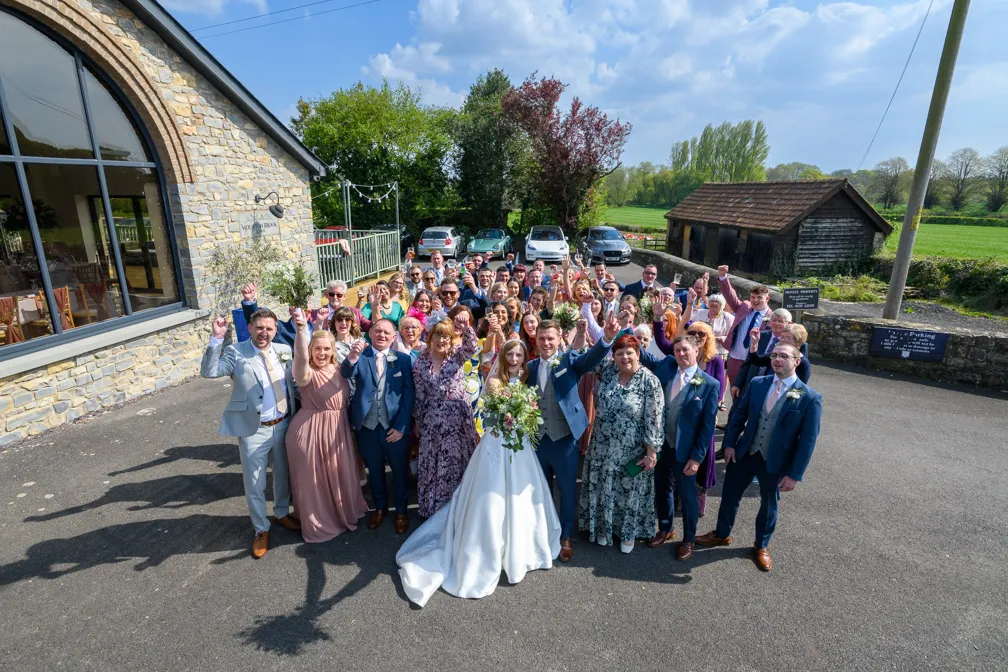 Large group of wedding guests posing together outdoors with the bride and groom in the center, outside a stone building on a sunny day.