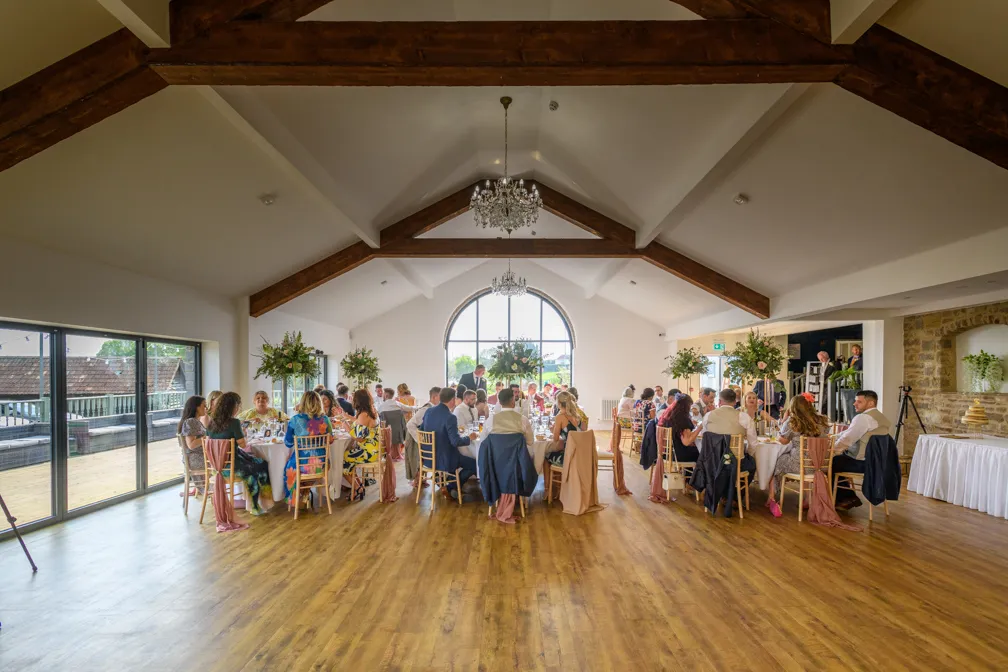 Guests seated at decorated round tables inside a bright event hall with wooden beams and chandeliers.