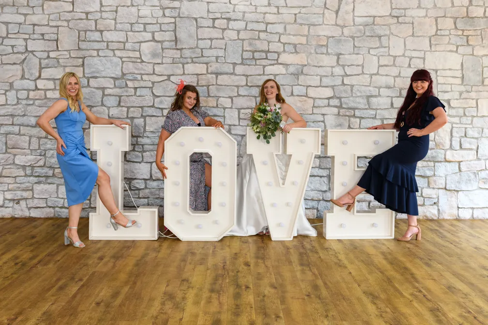 Four women in dresses posing with large illuminated letters spelling LOVE against a stone wall and wooden floor.