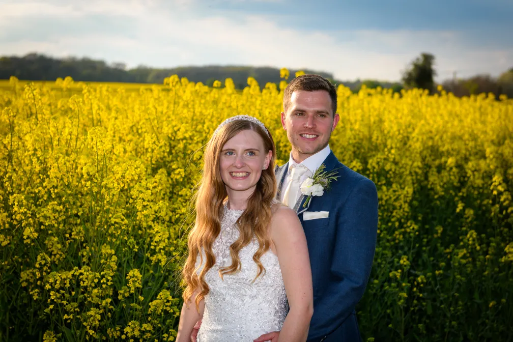 Bride with long wavy hair in a white lace dress and groom in a blue suit with a boutonniere standing in a yellow flower field.