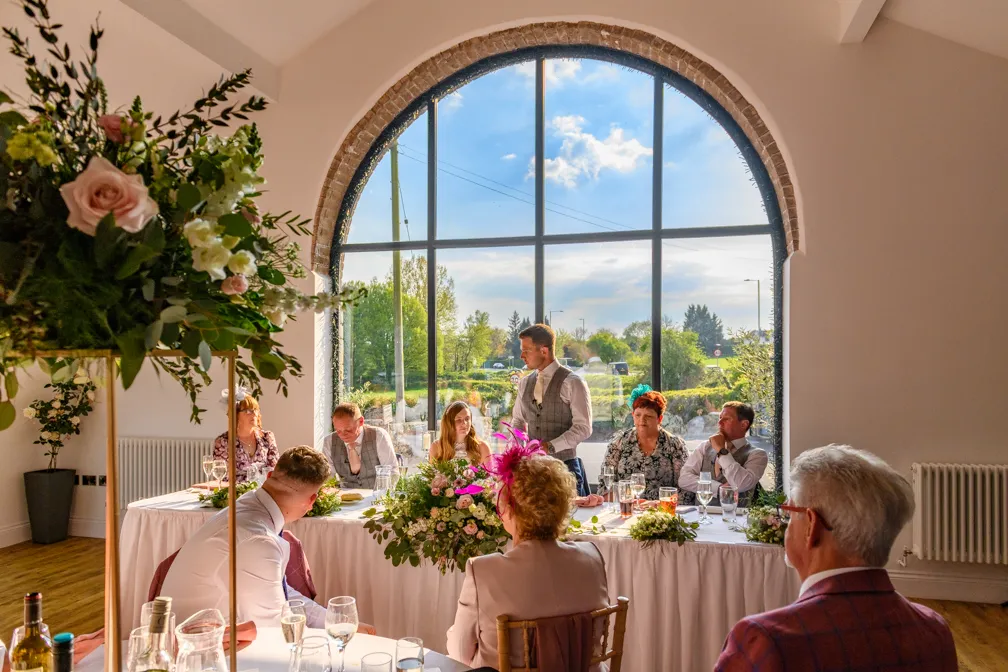 Man standing and speaking at a wedding head table with guests seated by a large arched window with outdoor greenery and blue sky visible.