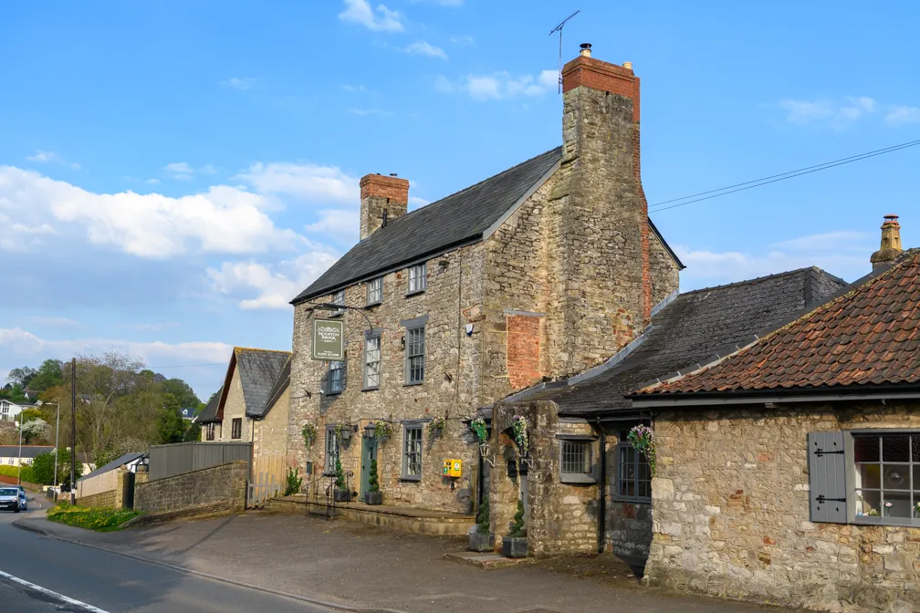 Stone building with two large chimneys beside a road under a partly cloudy blue sky.