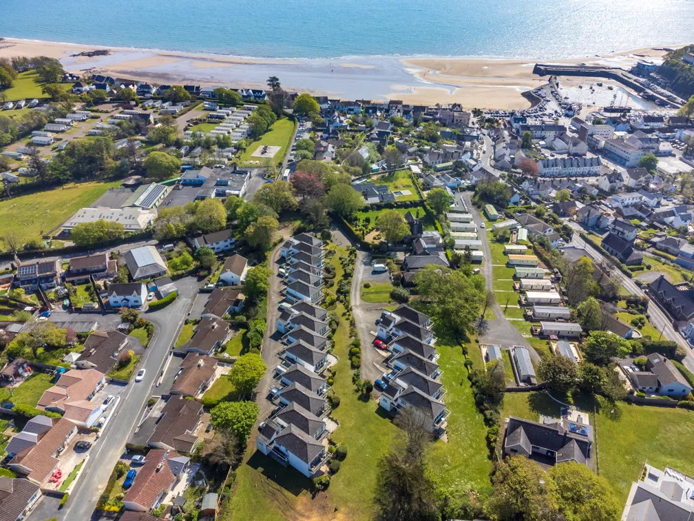 Aerial view of a coastal town with rows of houses, green lawns, and a sandy beach beside blue ocean water.